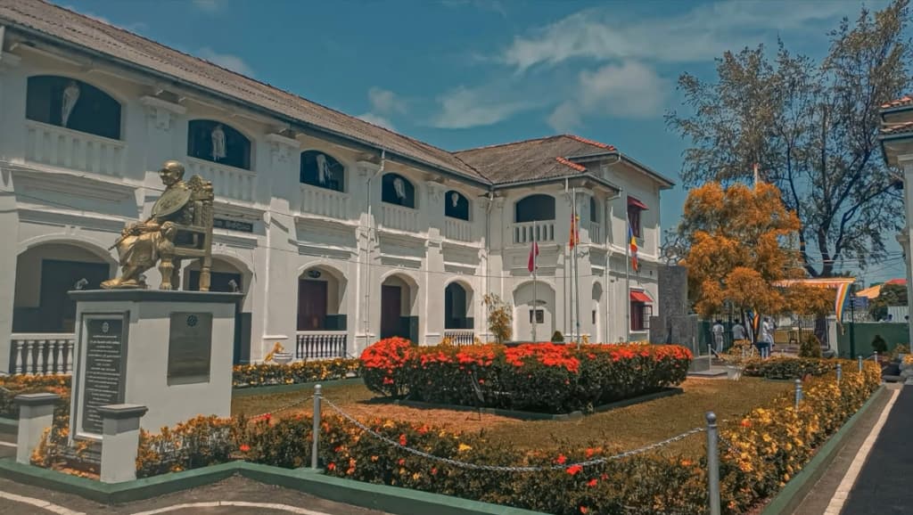 Seated statue of Anagarika Dharmapala in front of the white colonial-style Nalanda College building